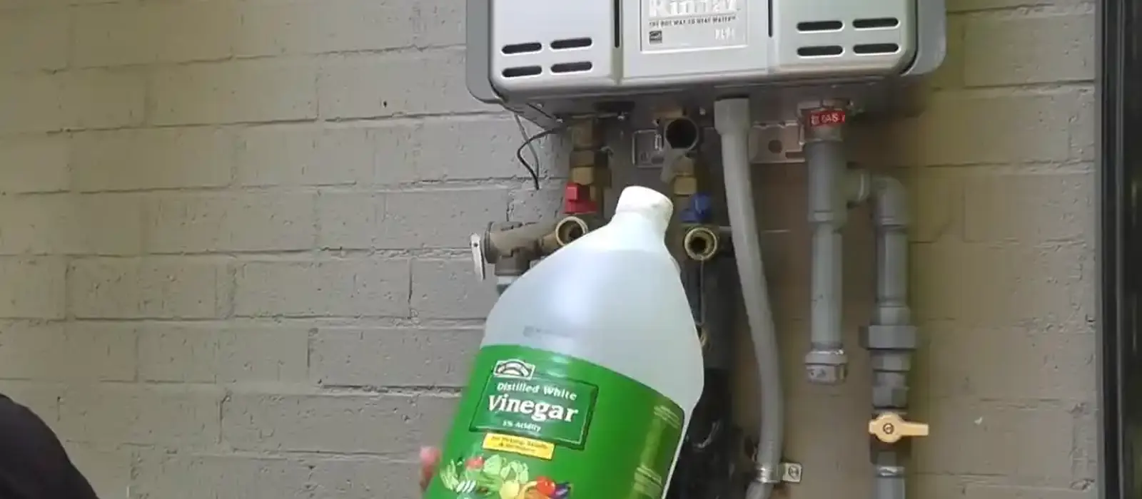 Jug of white vinegar held under a wall-mounted tankless water heater for a descaling flush.